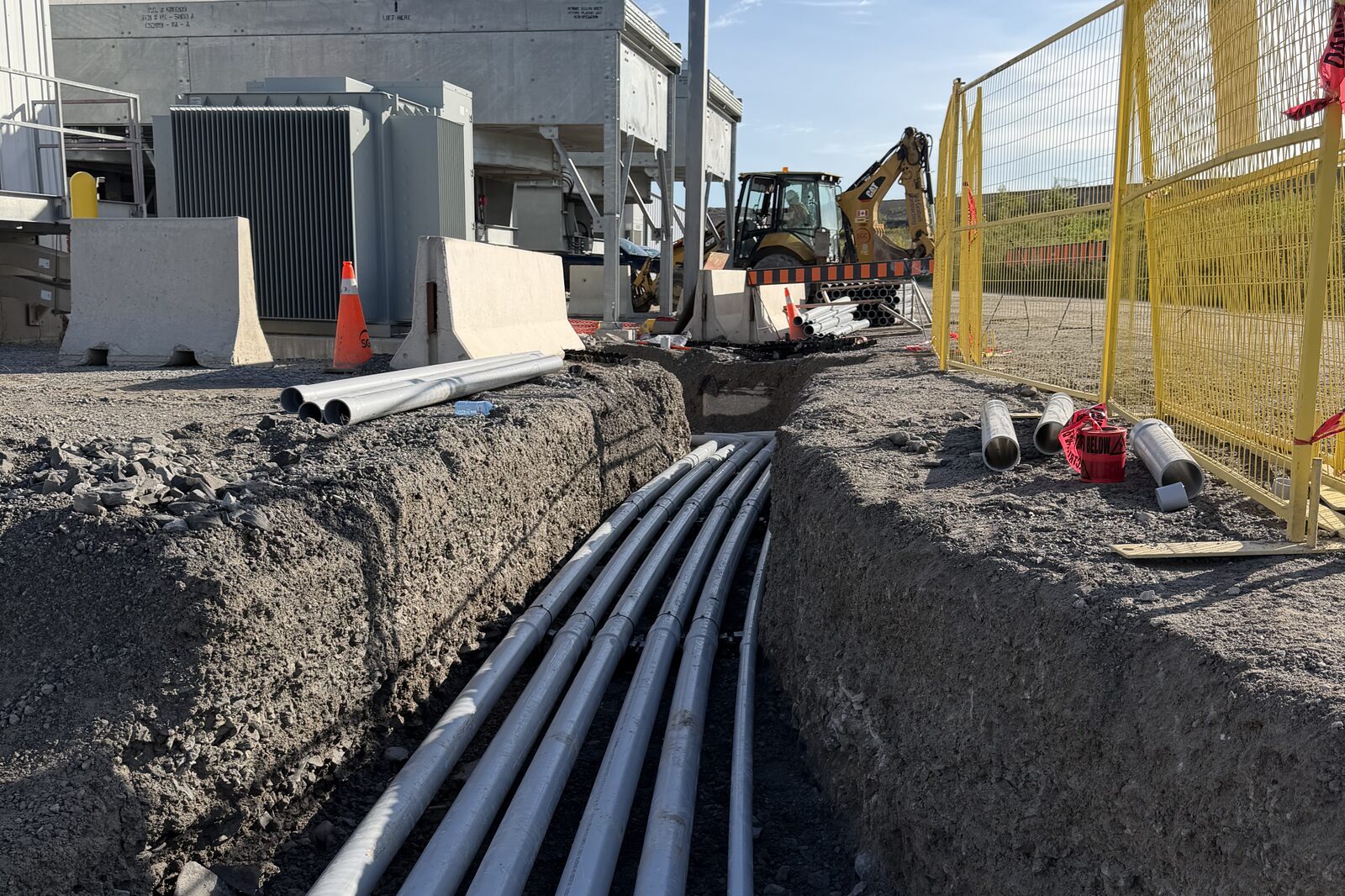 An open utility trench with bundled electrical conduit run through it, backhoe working in the background